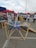 A group of people gathers at an outdoor event with tents in various colors. A geometric wooden structure is prominently displayed in the foreground, with two people interacting with it. The background shows other attendees and various activities taking place.