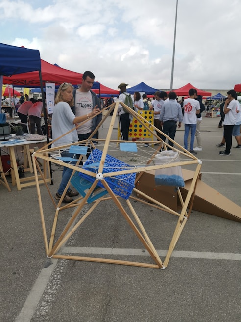 A group of people gathers at an outdoor event with tents in various colors. A geometric wooden structure is prominently displayed in the foreground, with two people interacting with it. The background shows other attendees and various activities taking place.