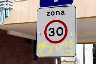A road sign displaying a speed limit of 30 is affixed to a pole. The sign is white with a red circle and black numbers. Above the sign, the word 'zona' is written. There is graffiti with a yellow tag over the lower part of the sign. A building with a white facade and a blue cloth is visible in the background.