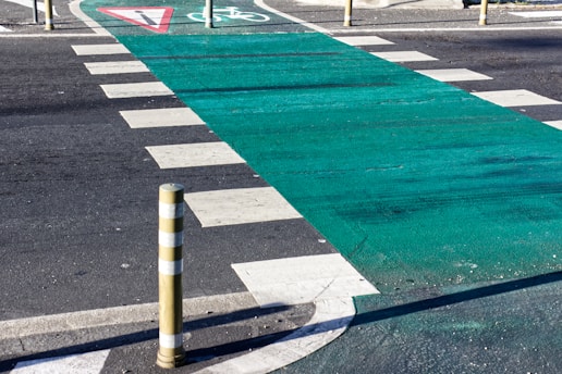 A crosswalk featuring a green bike lane running between white zebra stripes on a paved road. Yellow-striped bollards are positioned at intervals, while a triangular yield sign is visible on the ground.