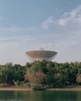 A large satellite dish stands prominently amidst a dense canopy of green trees. The structure has a lattice framework and is positioned near a serene body of water, with a clear blue sky overhead.