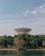 A large satellite dish stands prominently amidst a dense canopy of green trees. The structure has a lattice framework and is positioned near a serene body of water, with a clear blue sky overhead.
