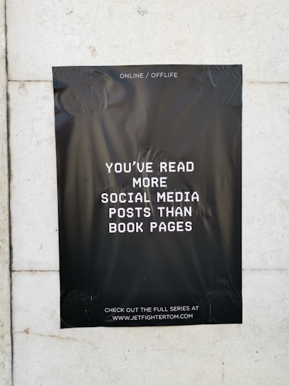A black poster with white text is taped to a light-colored wall. The text on the poster reads, 'You've read more social media posts than book pages.' The background of the poster is plain black, creating a stark contrast with the white lettering. The poster contains a website address at the bottom, inviting viewers to visit.
