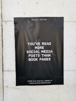 A black poster with white text is taped to a light-colored wall. The text on the poster reads, 'You've read more social media posts than book pages.' The background of the poster is plain black, creating a stark contrast with the white lettering. The poster contains a website address at the bottom, inviting viewers to visit.