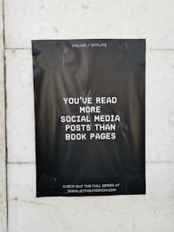 A black poster with white text is taped to a light-colored wall. The text on the poster reads, 'You've read more social media posts than book pages.' The background of the poster is plain black, creating a stark contrast with the white lettering. The poster contains a website address at the bottom, inviting viewers to visit.