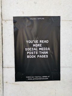 A black poster with white text is taped to a light-colored wall. The text on the poster reads, 'You've read more social media posts than book pages.' The background of the poster is plain black, creating a stark contrast with the white lettering. The poster contains a website address at the bottom, inviting viewers to visit.