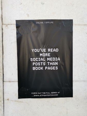 A black poster with white text is taped to a light-colored wall. The text on the poster reads, 'You've read more social media posts than book pages.' The background of the poster is plain black, creating a stark contrast with the white lettering. The poster contains a website address at the bottom, inviting viewers to visit.