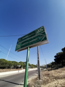A green road sign is positioned along a roadside, providing directions for cyclists to Trafaria and Costa da Caparica, with distances indicated in kilometers. The road is bordered by trees and a clear blue sky overhead. A lamp post and power lines are also visible, adding to the rural and serene setting.