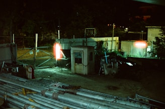 A dimly lit industrial area features a small structure surrounded by metal fencing. There are piles of pipes and construction materials scattered across the ground. Bright lights cast an eerie glow, illuminating parts of the scene, creating strong contrasts between light and shadow.