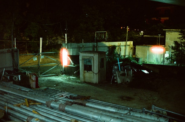 A dimly lit industrial area features a small structure surrounded by metal fencing. There are piles of pipes and construction materials scattered across the ground. Bright lights cast an eerie glow, illuminating parts of the scene, creating strong contrasts between light and shadow.