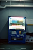 A dimly lit vending machine stands against a plain wall, stocked with rows of bottled beverages, including water and isotonic drinks. The machine is predominantly blue with illuminated sections highlighting the available products and a signage that reads 'Bonaqua BonACTIVE Isotonic'.