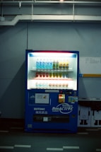 A dimly lit vending machine stands against a plain wall, stocked with rows of bottled beverages, including water and isotonic drinks. The machine is predominantly blue with illuminated sections highlighting the available products and a signage that reads 'Bonaqua BonACTIVE Isotonic'.