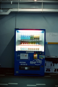A dimly lit vending machine stands against a plain wall, stocked with rows of bottled beverages, including water and isotonic drinks. The machine is predominantly blue with illuminated sections highlighting the available products and a signage that reads 'Bonaqua BonACTIVE Isotonic'.