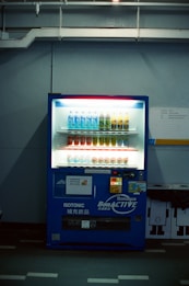 A dimly lit vending machine stands against a plain wall, stocked with rows of bottled beverages, including water and isotonic drinks. The machine is predominantly blue with illuminated sections highlighting the available products and a signage that reads 'Bonaqua BonACTIVE Isotonic'.