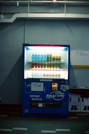 A dimly lit vending machine stands against a plain wall, stocked with rows of bottled beverages, including water and isotonic drinks. The machine is predominantly blue with illuminated sections highlighting the available products and a signage that reads 'Bonaqua BonACTIVE Isotonic'.