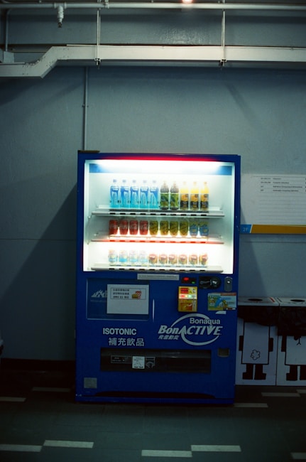 A dimly lit vending machine stands against a plain wall, stocked with rows of bottled beverages, including water and isotonic drinks. The machine is predominantly blue with illuminated sections highlighting the available products and a signage that reads 'Bonaqua BonACTIVE Isotonic'.