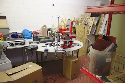 A cluttered workshop or storage room with various tools and equipment placed on tables and shelves. The room is filled with different materials, including boxes, plastic containers, and wooden boards. A large machine with a red component is prominently placed on a round table in the center. The room has white tiled walls and a red tiled floor.