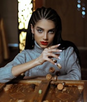 A person with braided hair and striking makeup intently focuses on a backgammon game, holding several game pieces mid-air. The setting is indoors, with warm and dim lighting, contributing to an intimate atmosphere. The person wears a gray turtleneck sweater.