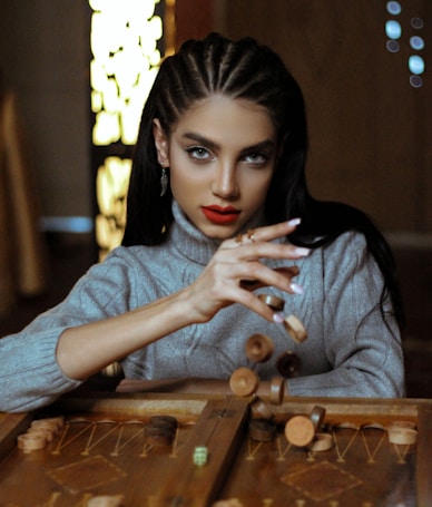 A person with braided hair and striking makeup intently focuses on a backgammon game, holding several game pieces mid-air. The setting is indoors, with warm and dim lighting, contributing to an intimate atmosphere. The person wears a gray turtleneck sweater.