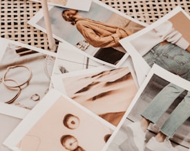 A collection of photographs scattered on a wicker surface. The images depict various subjects, including a person wearing a brown sweater, a pair of jeans with white sneakers, several pieces of jewelry like rings and earrings, and abstract close-ups of fabric and apples. The overall tone appears warm and vintage.