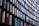 A modern architectural facade with a repetitive pattern of vertical panels. The panels have reflective surfaces and are arranged in a curved design, displaying a mix of red, blue, and neutral colors. The design creates a dynamic and visually striking appearance.