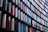 A modern architectural facade with a repetitive pattern of vertical panels. The panels have reflective surfaces and are arranged in a curved design, displaying a mix of red, blue, and neutral colors. The design creates a dynamic and visually striking appearance.