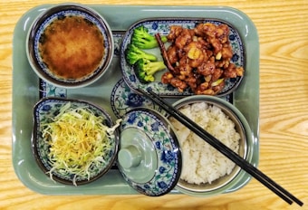 A variety of dishes served on a green tray consisting of a bowl of soup, a side of stir-fried vegetables with meat, steamed rice, and a salad featuring shredded cabbage. The dishes are styled in traditional Asian ceramic ware, adorned with intricate blue and floral patterns. Chopsticks are placed across the dish with rice.