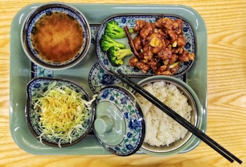 A variety of dishes served on a green tray consisting of a bowl of soup, a side of stir-fried vegetables with meat, steamed rice, and a salad featuring shredded cabbage. The dishes are styled in traditional Asian ceramic ware, adorned with intricate blue and floral patterns. Chopsticks are placed across the dish with rice.