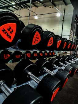 A row of black dumbbells with orange labels indicating various weights is neatly arranged on a rack in a gym setting. The dumbbells are reflected in a mirror behind them, and the gym has a visible ceiling structure with overhead lights.