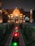A miniature replica of the Taj Mahal is illuminated with warm yellow and orange lights, set against a dark evening sky. The structure is surrounded by lush greenery, and a channel of water with green and red illuminated fountains leads up to the building.