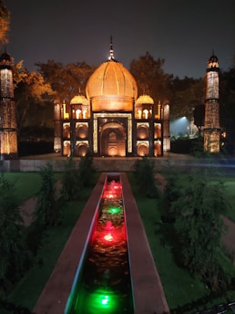 A miniature replica of the Taj Mahal is illuminated with warm yellow and orange lights, set against a dark evening sky. The structure is surrounded by lush greenery, and a channel of water with green and red illuminated fountains leads up to the building.