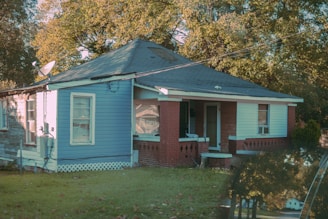 A small, single-story house painted in blue with a red brick porch, surrounded by green grass and leafy trees. The house has a slanted roof and multiple windows, some of which have air conditioning units. A satellite dish is attached to the side of the house, and fallen leaves are scattered on the lawn.