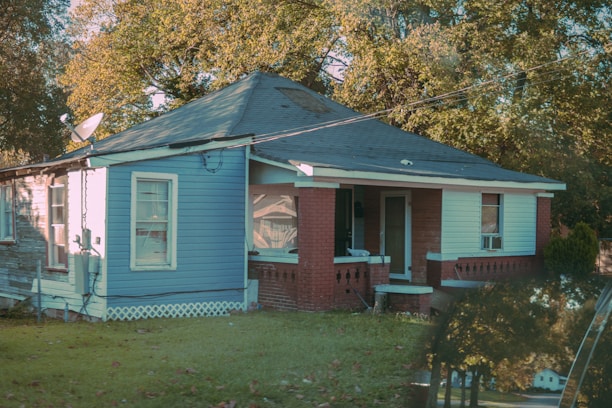 A small, single-story house painted in blue with a red brick porch, surrounded by green grass and leafy trees. The house has a slanted roof and multiple windows, some of which have air conditioning units. A satellite dish is attached to the side of the house, and fallen leaves are scattered on the lawn.