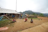 A rural playground scene shows children playing on various equipment set on dirt ground. A metal slide is occupied by two children while others play on a see-saw and other apparatus. A simple building with a corrugated metal roof is in the background. Clothes are hanging to dry, and there are mountains partially covered by mist in the distance.