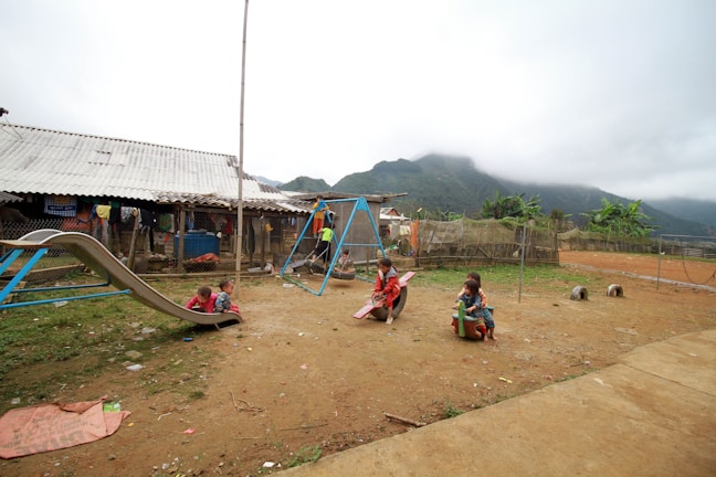 A rural playground scene shows children playing on various equipment set on dirt ground. A metal slide is occupied by two children while others play on a see-saw and other apparatus. A simple building with a corrugated metal roof is in the background. Clothes are hanging to dry, and there are mountains partially covered by mist in the distance.