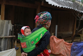 woman in black long-sleeved shirt carrying her child while walking beside brown wooden house during daytime