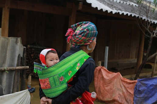 woman in black long-sleeved shirt carrying her child while walking beside brown wooden house during daytime