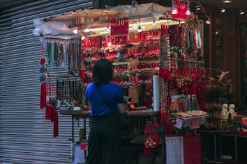 A person with a blue shirt stands in front of a vibrant street market stall adorned with a wide array of colorful trinkets and hanging accessories. The stall is brightly illuminated, contrasting with the surrounding dim environment, and features numerous red decorations and beaded items.