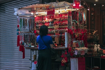 A person with a blue shirt stands in front of a vibrant street market stall adorned with a wide array of colorful trinkets and hanging accessories. The stall is brightly illuminated, contrasting with the surrounding dim environment, and features numerous red decorations and beaded items.