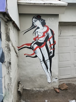 A mural painted on an outdoor wall depicts a stylized image of a woman with long flowing black hair. Her body is wrapped in vibrant red ribbons that accentuate the monochromatic black and white tones of her figure. The artwork has a dynamic and fluid composition set against a rough, textured concrete wall. Graffiti tags are visible beside the figure.