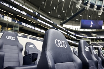 A row of black leather stadium seats with the Audi logo prominently displayed in an upscale sports venue. In the background, there are large screens and bright signage displaying the words 'The game is about glory,' suggesting a high-profile sporting event location.