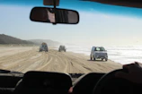 4x4 vehicle driving along a sandy beach with kitesurfers preparing their gear nearby.