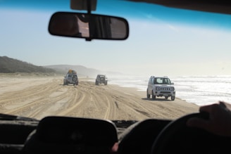 A convoy of 4x4 vehicles driving along a coastal road near the Emerald Sea with turquoise waters visible.