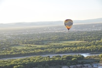A colorful hot air balloon hovers above a sprawling landscape filled with lush green trees. The scene captures a broad view of a city with mountains in the background, under a clear sky. The sunlight casts a warm glow over the area, creating a serene and picturesque atmosphere.