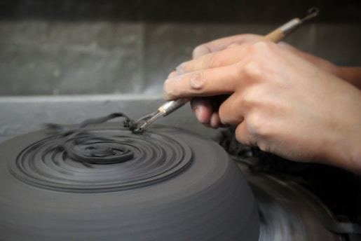 Hands are crafting a circular clay object on a spinning pottery wheel, using a small tool to shape and detail the edges. The workspace is a muted, earthy tone.