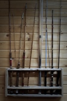 Several fishing rods are neatly organized on a wooden rack mounted against a wooden wall. The rods vary in color and style, some with red or black accents, alongside wood and metallic finishes.