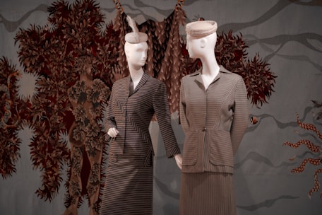 Two mannequins dressed in vintage-style clothing, featuring striped patterns, are positioned against a textured backdrop with intricate, earth-toned foliage designs. One mannequin wears a hat adorned with a feather, adding a touch of elegance.