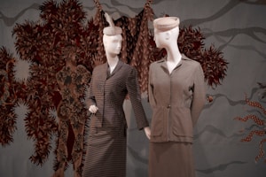 Two mannequins dressed in vintage-style clothing, featuring striped patterns, are positioned against a textured backdrop with intricate, earth-toned foliage designs. One mannequin wears a hat adorned with a feather, adding a touch of elegance.