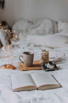 An open book lies on a soft white bed, creating a cozy and inviting atmosphere. Next to it, a wooden tray holds a ceramic mug and a vintage camera, suggesting a scene of relaxation and leisure. Twinkling fairy lights add a warm and soft glow to the background.