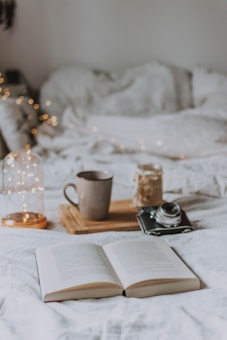 An open book lies on a soft white bed, creating a cozy and inviting atmosphere. Next to it, a wooden tray holds a ceramic mug and a vintage camera, suggesting a scene of relaxation and leisure. Twinkling fairy lights add a warm and soft glow to the background.